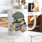 A white ceramic coffee mug with a cute sloth illustration wearing a green beanie and holding a coffee cup and a pink phone, set on a rustic white wooden table with a blurred kitchen background. Grab your mug from Coffee and moo NZ.