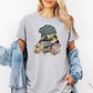 A woman wearing a heather gray oversized t-shirt with a cartoon graphic of a pug wearing a green beanie and holding a coffee cup and a phone. She has a light blue denim jacket draped off her shoulders against a plain white background. Grab your tee from Coffee and Moo NZ.