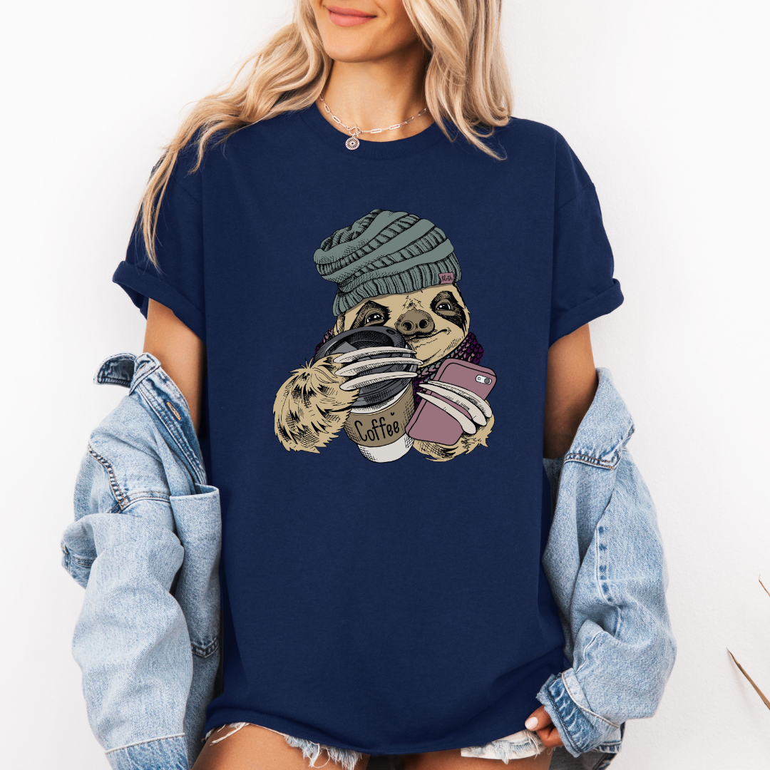 A woman wearing a navy blue oversized graphic t-shirt with a sloth illustration holding coffee cups, paired with a light blue denim jacket draped off her shoulders, against a plain white background. Grab your tee from Coffee and Moo NZ.