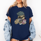 A woman wearing a navy blue oversized graphic t-shirt with a sloth illustration holding coffee cups, paired with a light blue denim jacket draped off her shoulders, against a plain white background. Grab your tee from Coffee and Moo NZ.