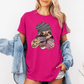 A woman wearing a bright pink graphic t-shirt with a cartoon sloth holding a coffee cup, styled with an open light blue denim jacket. Grab your tee from Coffee and Moo NZ.