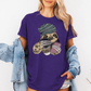 A woman wearing a purple short-sleeve t-shirt with a cartoon graphic of a pug wearing a green beanie and holding a coffee cup and a phone. She has a light blue denim jacket draped off her shoulders against a plain white background. Grab your tee from Coffee and Moo NZ.
