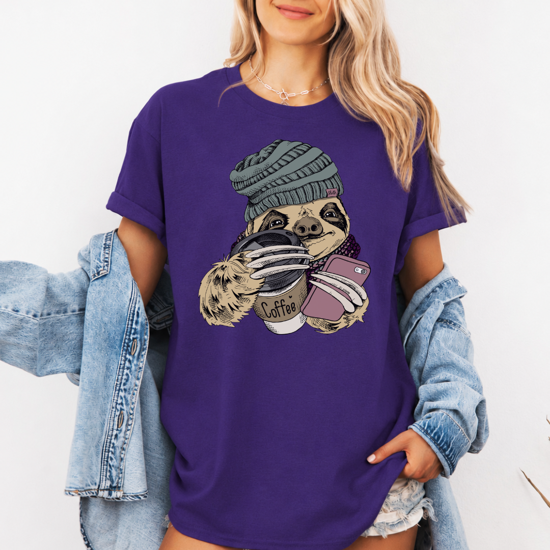 A woman wearing a purple short-sleeve t-shirt with a cartoon graphic of a pug wearing a green beanie and holding a coffee cup and a phone. She has a light blue denim jacket draped off her shoulders against a plain white background. Grab your tee from Coffee and Moo NZ.