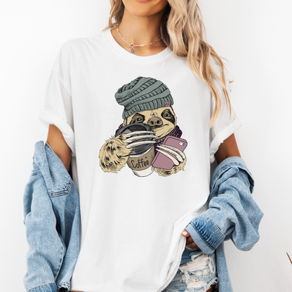 A woman wearing a white graphic t-shirt with a cartoon sloth holding a coffee cup and wearing a beanie. She has a light blue denim jacket off her shoulders and is posing against a plain white background. Grab your tee from Coffee and Moo NZ.