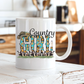 A white ceramic coffee mug with a 'Country Girl' graphic design, sitting on a rustic white wooden table with a blurred kitchen background.  From Coffee and Moo NZ