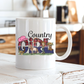 A white ceramic coffee mug with a 'Country Girl' graphic print, sitting on a rustic white wooden table with a blurred kitchen background. From Coffee and Moo NZ.