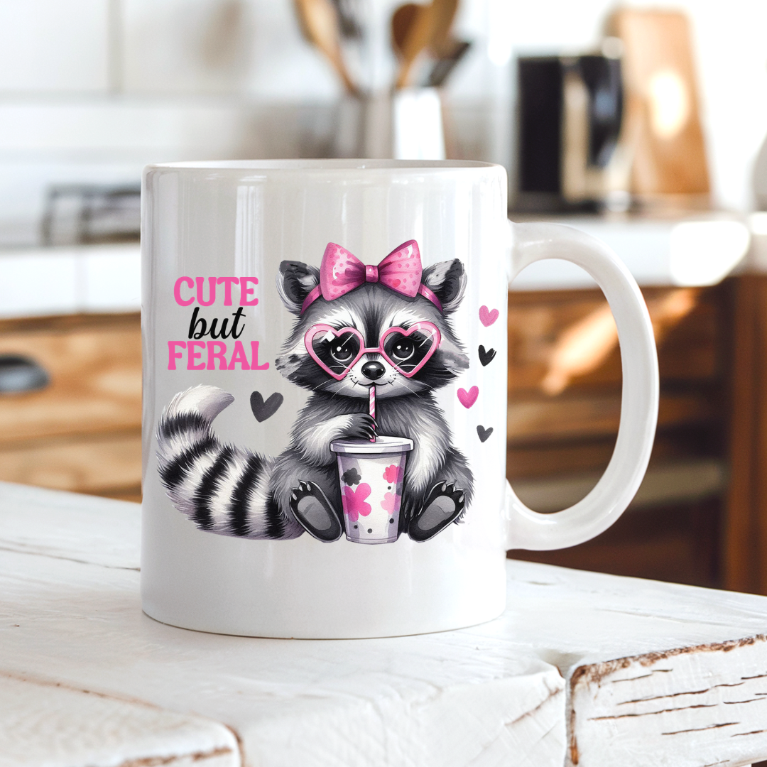 A white ceramic coffee mug with a cartoon raccoon wearing a pink bow and heart glasses, holding a drink, with the text 'CUTE but FERAL' in pink and black. The mug is sitting on a rustic white wooden table with a blurred kitchen background. from Coffee and Moo.
