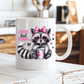 A white ceramic coffee mug with a cartoon raccoon wearing a pink bow and heart glasses, holding a drink, with the text 'CUTE but FERAL' in pink and black. The mug is sitting on a rustic white wooden table with a blurred kitchen background. from Coffee and Moo.