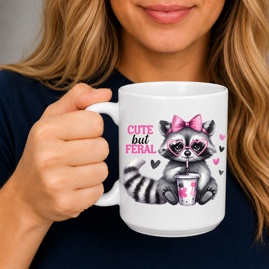 A person holding a white ceramic mug with a cute cartoon raccoon holding a drink and the text 'CUTE but FERAL' in pink and black, wearing a navy shirt. from Coffee and Moo.
