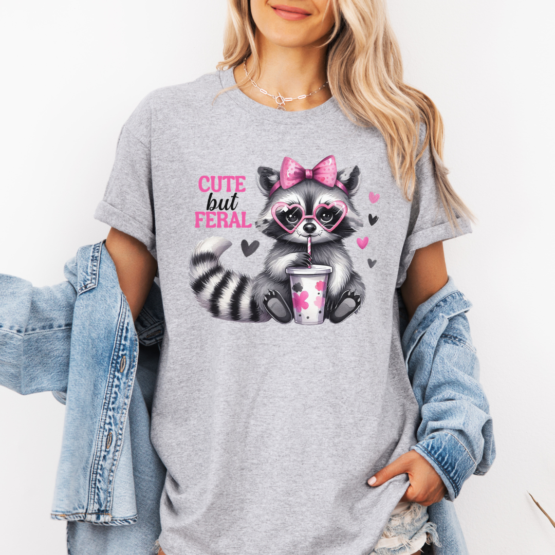 A woman wearing a grey graphic t-shirt with a cartoon raccoon holding a pink drink and the text 'CUTE but FERAL'. She has a light blue denim jacket draped off her shoulders against a plain white background. purchase from Coffee and Moo NZ.