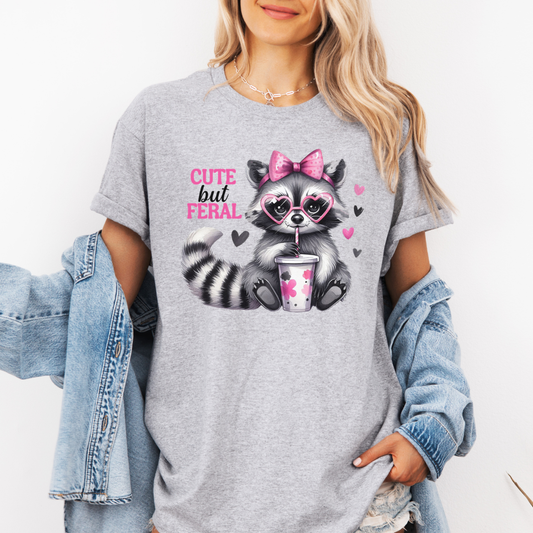A woman wearing a grey graphic t-shirt with a cartoon raccoon holding a pink drink and the text 'CUTE but FERAL'. She has a light blue denim jacket draped off her shoulders against a plain white background. purchase from Coffee and Moo NZ.