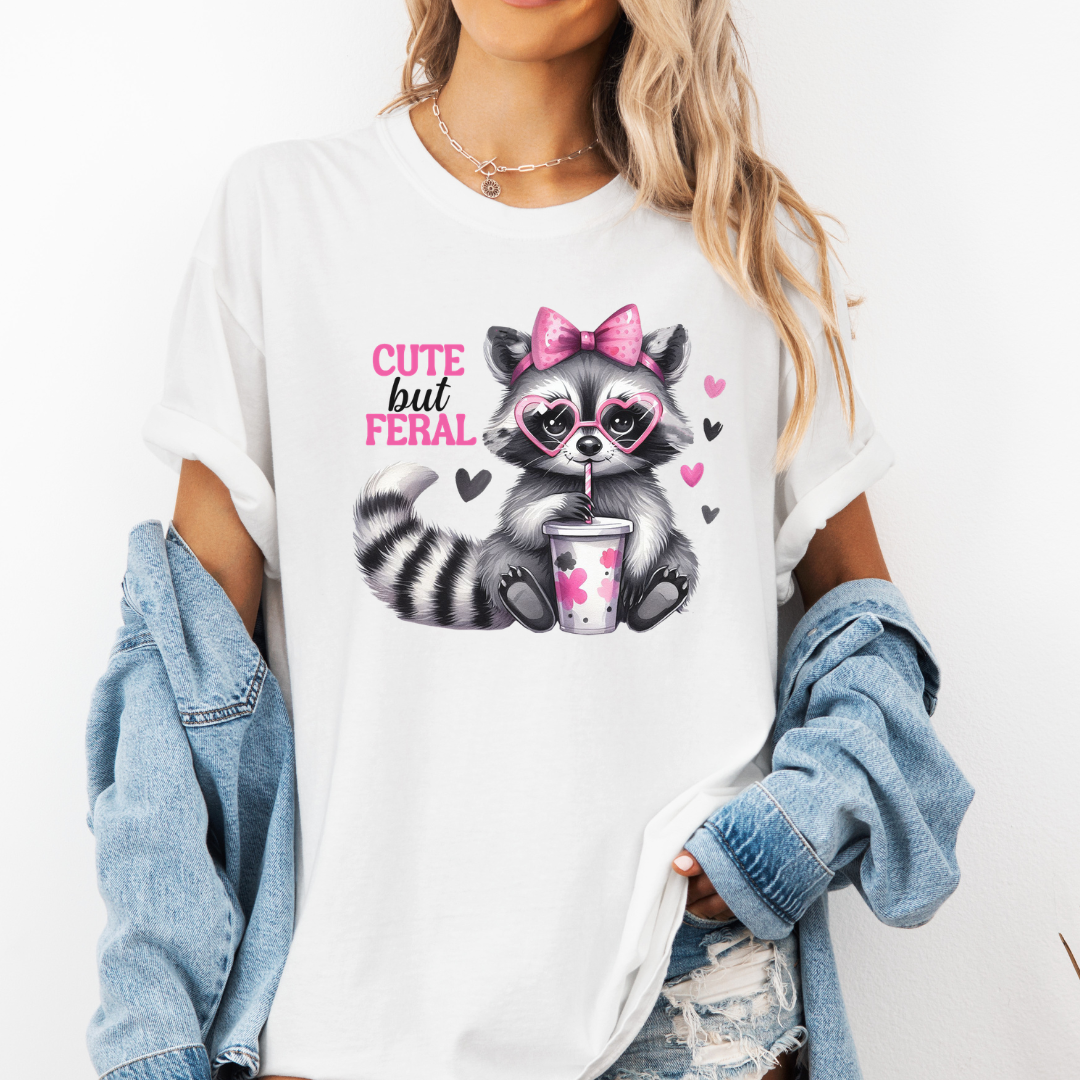 A woman wearing a white graphic t-shirt with a cute raccoon illustration and the text 'CUTE but FERAL'. She has a light blue denim jacket draped off her shoulders and is wearing distressed denim shorts against a plain white background. purchase from Coffee and Moo NZ.
