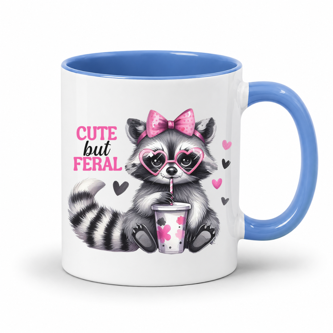 A white ceramic mug with a blue interior and handle, featuring a cute cartoon raccoon wearing pink heart glasses and a pink bow, holding a drink cup, with the text 'CUTE but FERAL' on the front. from Coffee and Moo.