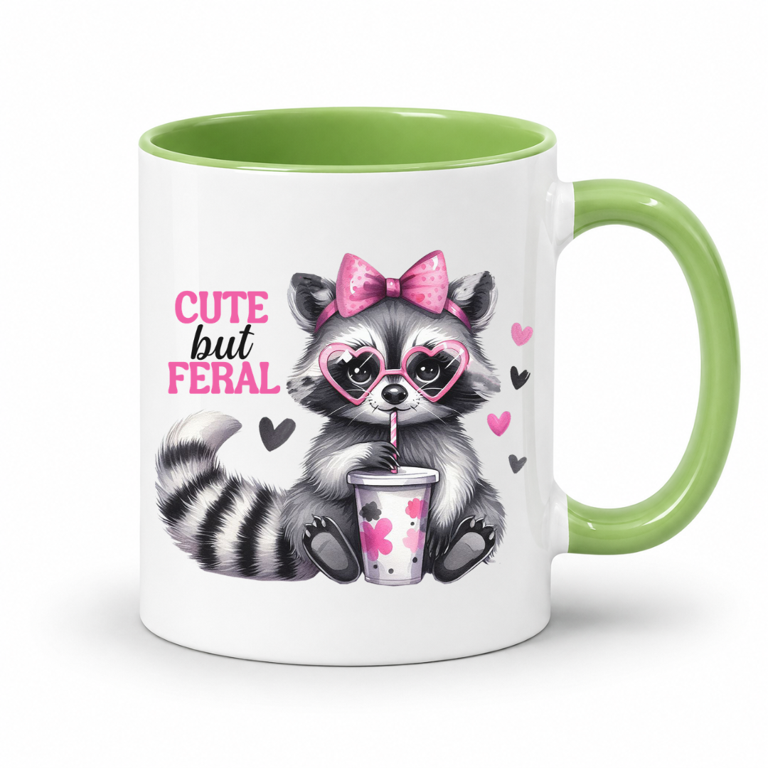 A white ceramic mug with a light green interior and handle, featuring a cute cartoon raccoon wearing pink heart glasses and a pink bow, holding a drink cup, with the text 'CUTE but FERAL' printed on the side. from Coffee and Moo.