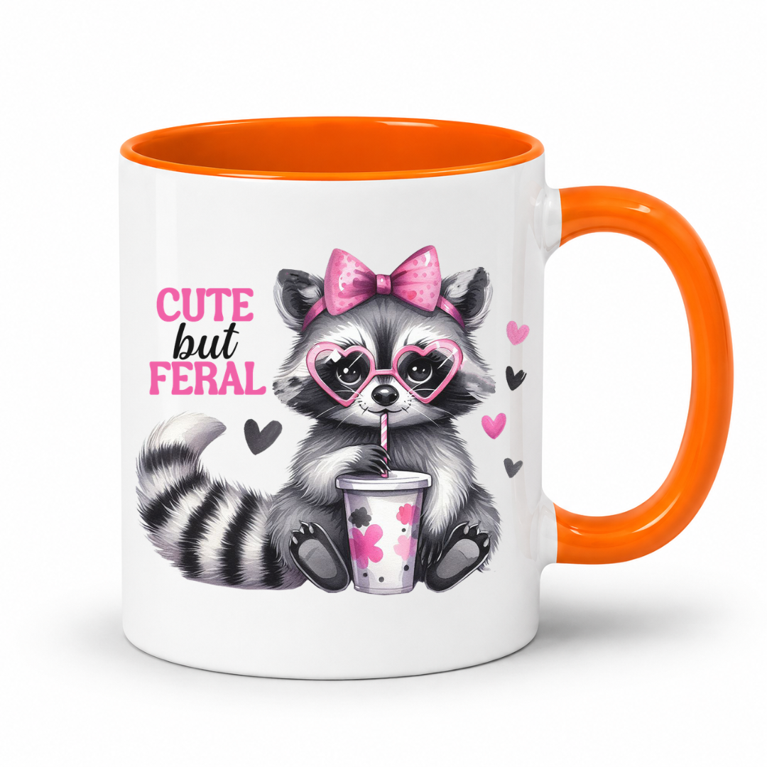 A white ceramic mug with a bright orange handle and interior, featuring a cute cartoon raccoon wearing pink heart-shaped sunglasses and a pink bow, holding a drink, with the text 'CUTE but FERAL' on the front. from Coffee and Moo.