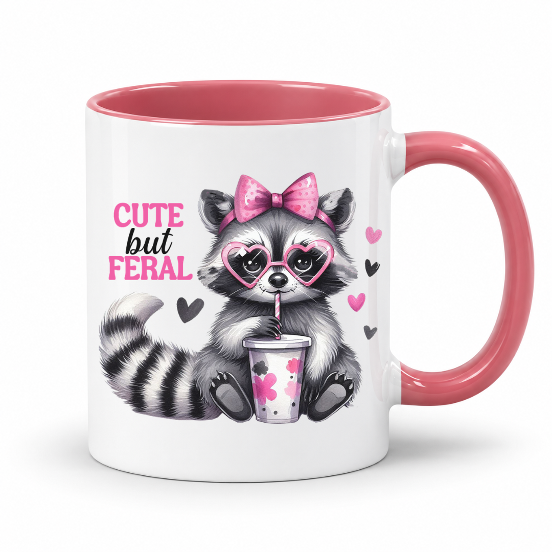 A white ceramic mug with a pink interior and handle, featuring a cute cartoon raccoon wearing heart-shaped sunglasses and a pink bow, holding a drink cup, with the text 'CUTE but FERAL' printed on the side. from Coffee and Moo.