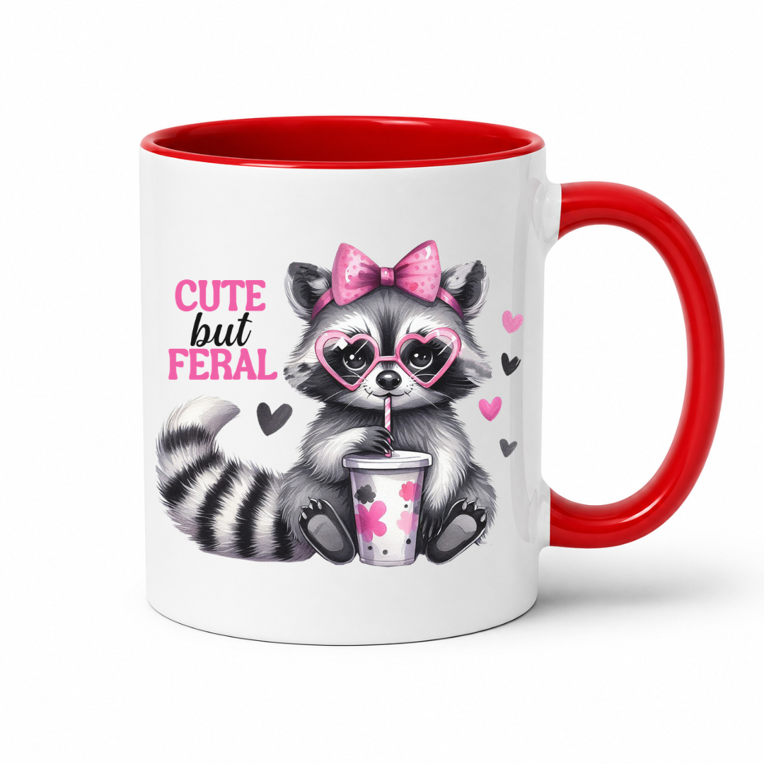 A white ceramic mug with a red interior, red handle, and a printed graphic of a raccoon wearing heart-shaped glasses and a pink bow, holding a drink, with the text 'CUTE but FERAL'. from Coffee and Moo.