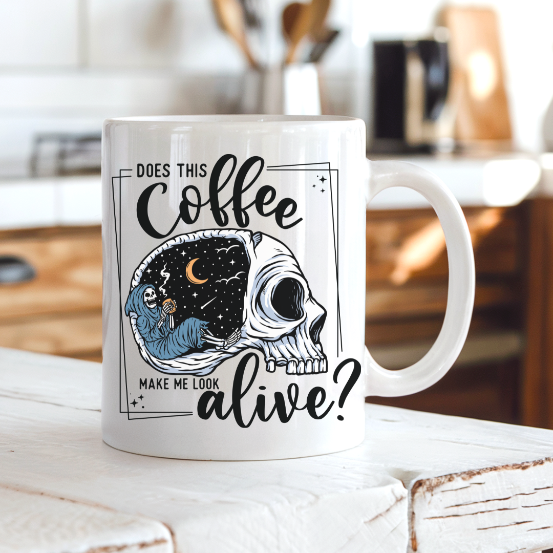 A white ceramic coffee mug with a skull graphic design, sitting on a rustic white wooden table in a warm, blurred kitchen setting. The graphic features a skull with one half showing a starry night sky with a crescent moon and a skeleton holding a coffee, accompanied by the text 'DOES THIS Coffee MAKE ME LOOK alive?'. Grab yours from Coffee and Moo NZ.