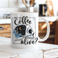 A white ceramic coffee mug with a skull graphic design, sitting on a rustic white wooden table in a warm, blurred kitchen setting. The graphic features a skull with one half showing a starry night sky with a crescent moon and a skeleton holding a coffee, accompanied by the text 'DOES THIS Coffee MAKE ME LOOK alive?'. Grab yours from Coffee and Moo NZ.