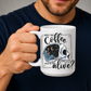 A man in a navy blue t-shirt holds a white ceramic coffee mug with a graphic that features a skull with one half showing a starry night sky with a crescent moon and a skeleton holding a coffee, accompanied by the text 'DOES THIS Coffee MAKE ME LOOK alive?'. Grab yours from Coffee and Moo NZ.