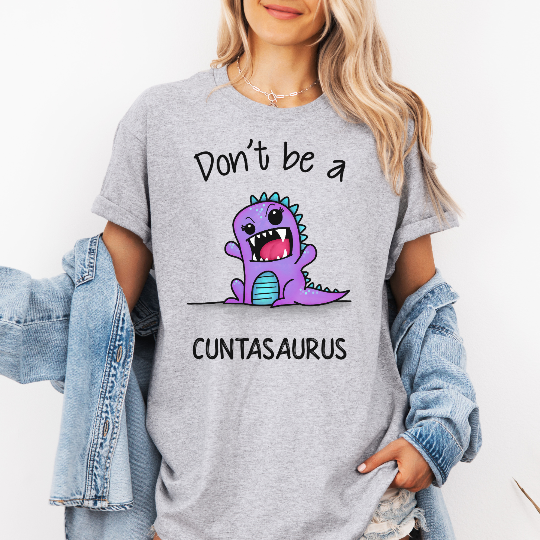 A woman wearing a grey graphic t-shirt with a purple cartoon dinosaur and the text 'Don't be a CUNTSAURUS'. She has a light blue denim jacket draped off her shoulders against a plain white background. Purchase from Coffee and Moo NZ.