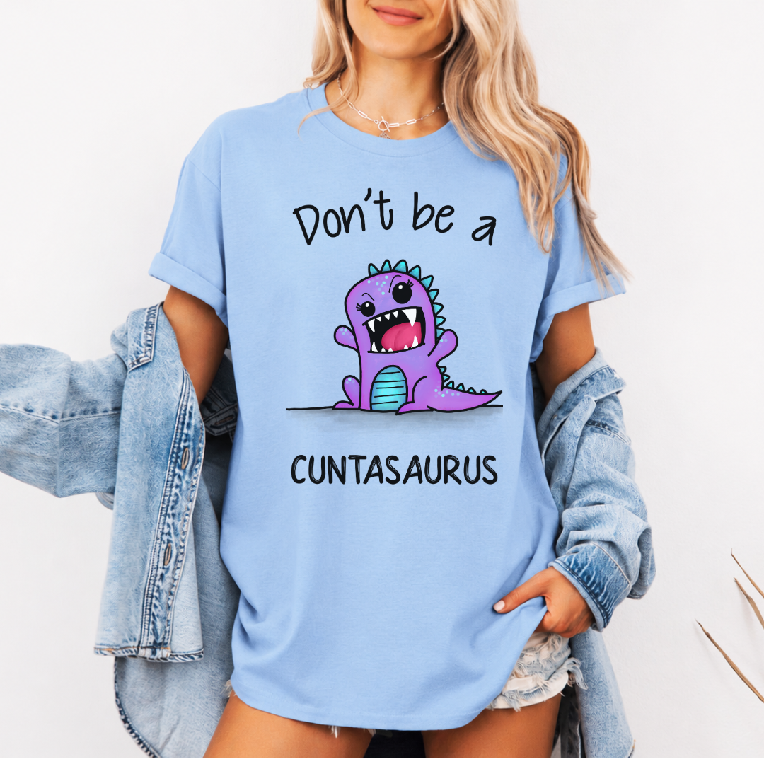 A woman wearing an oversized light blue graphic T-shirt with a purple cartoon dinosaur and the text 'Don't be a CUNTASaurus'. She has a light-wash denim jacket off her shoulders and is posing against a clean white background. Purchase from Coffee and Moo NZ.