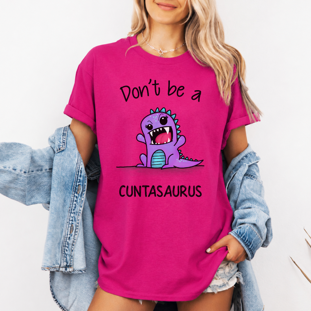 A woman wearing a bright pink graphic t-shirt with a purple cartoon dinosaur and the text 'Don't be a CUNTASaurus'. She has a light blue denim jacket off her shoulders against a plain white background. Purchase from Coffee and Moo NZ.