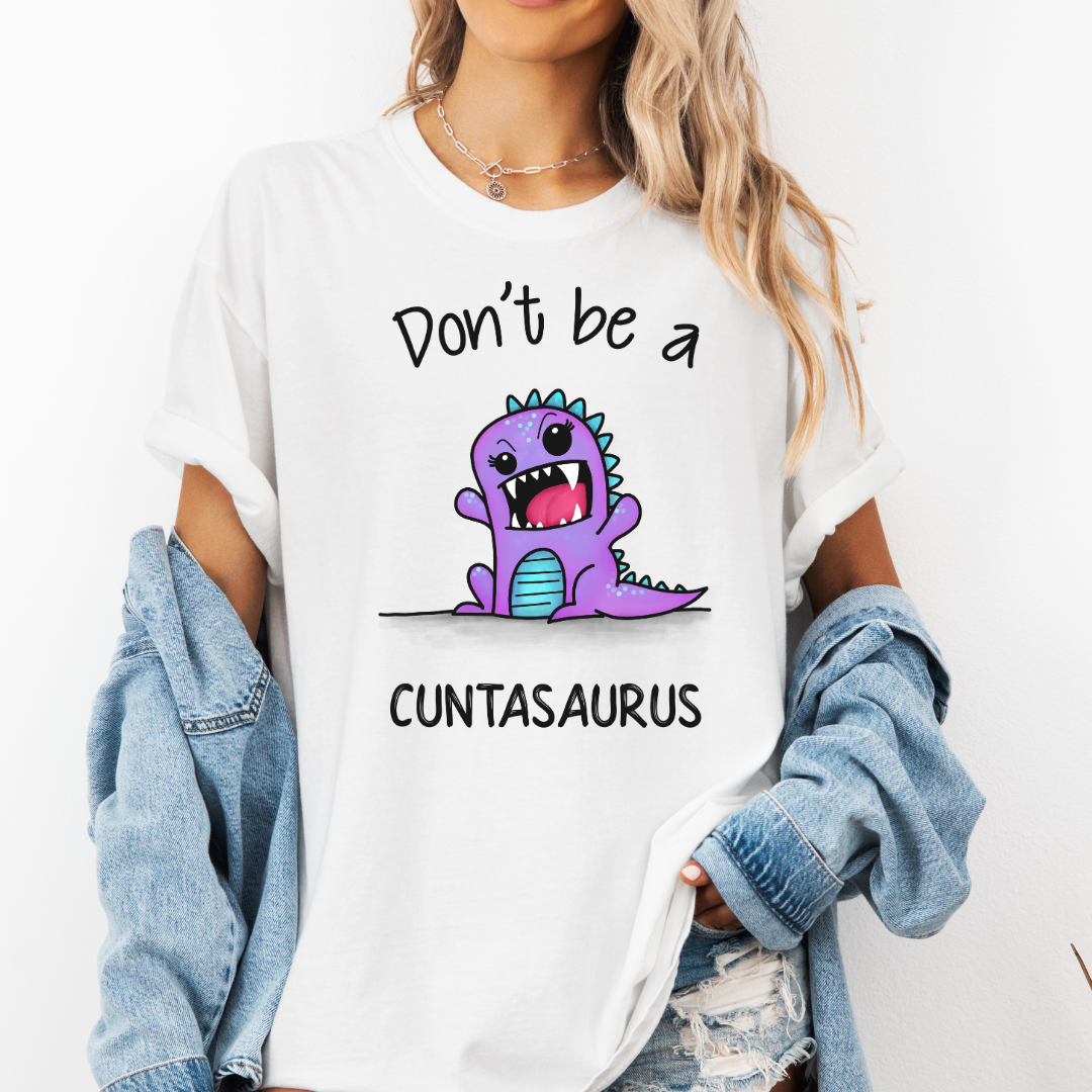 A woman wearing a white graphic t-shirt with a purple cartoon dinosaur and the text 'Don't be a CUNTASaurus'. She has a light blue denim jacket off her shoulders and is posing against a plain white wall. Purchase from Coffee and Moo NZ.
