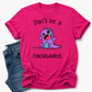 A bright pink graphic t-shirt featuring a cute purple cartoon dinosaur and the text 'Don't be a CUNTASaurus', styled flat-lay next to a pair of blue jeans on a white background. Grab your tee from Coffee and Moo NZ.