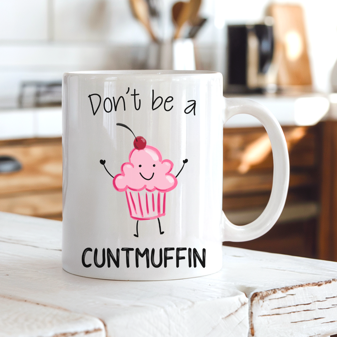 A white ceramic mug with a humorous print that reads 'Don't be a CUNTMUFFIN' in black text, featuring a cute pink cupcake character with a cherry on top, set on a rustic white wooden table with a blurred kitchen background. Grab yours from Coffee and Moo NZ.