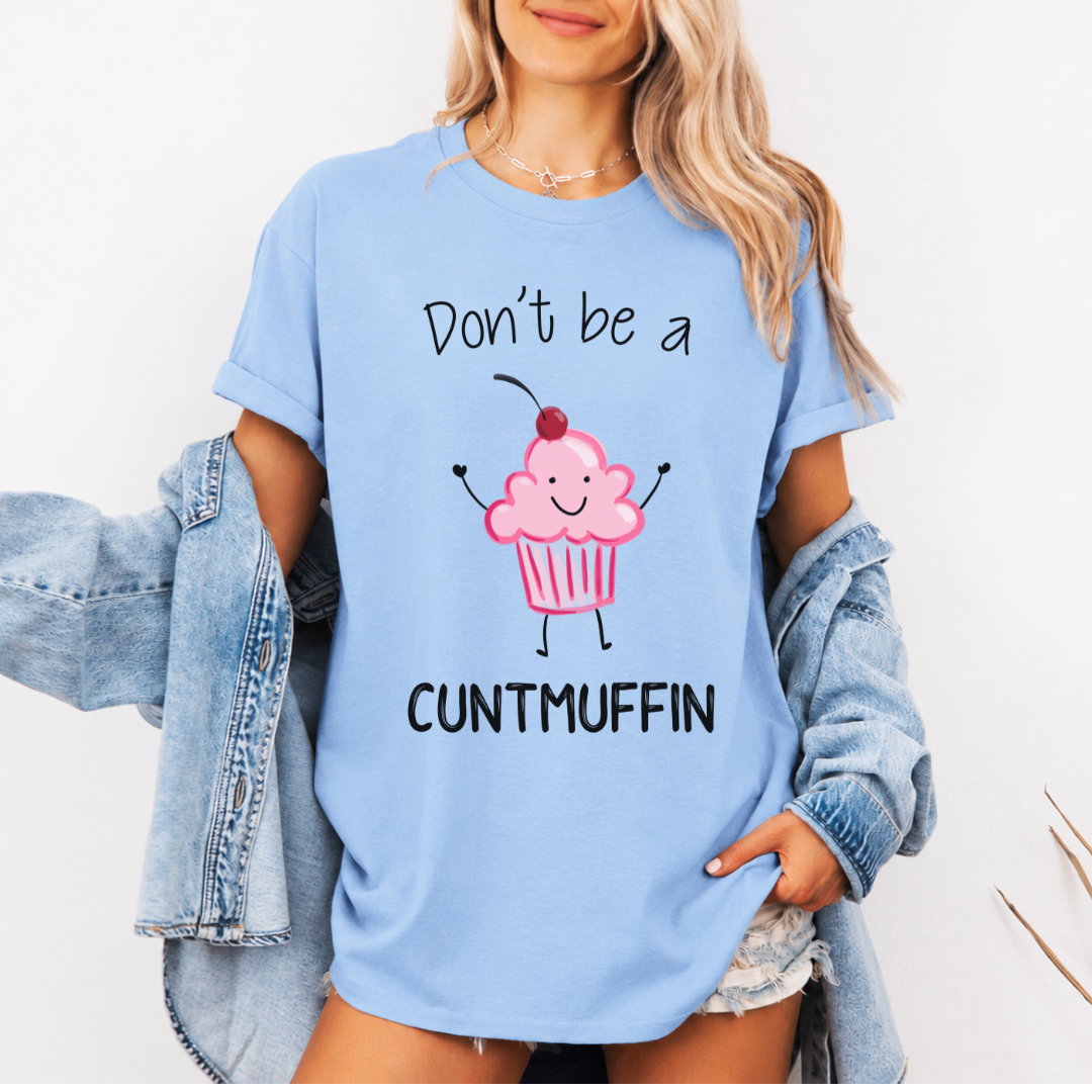 A woman wearing a light blue graphic t-shirt with a pink cupcake character and the text 'Don't be a CUNTMUFFIN'. She has a light wash denim jacket off her shoulders and is posing against a plain white background. Purchase from Coffee and Moo NZ.