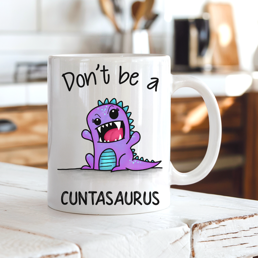 A white ceramic coffee mug with a cartoon purple dinosaur and the text 'Don't be a CUNTASaurus' printed on it, set on a rustic white wooden table with a blurred kitchen background. Grab yours from Coffee and Moo NZ.
