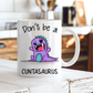 A white ceramic coffee mug with a cartoon purple dinosaur and the text 'Don't be a CUNTASaurus' printed on it, set on a rustic white wooden table with a blurred kitchen background. Grab yours from Coffee and Moo NZ.