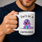 A man holding a white ceramic mug with a cartoon purple dinosaur graphic and the text 'Don't be a CUNTASaurus'. Grab yours from Coffee and Moo.