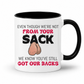 A white ceramic coffee mug with a black interior and handle, has a humorous graphic. The mug features the text 'EVEN THOUGH WE'RE NOT FROM YOUR SACK WE KNOW YOU'VE STILL GOT OUR BACKS' in black and red lettering, accompanied by a cartoon drawing of a hairy balls. Grab your mug from Coffee and Moo NZ.
