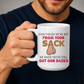 A man in a navy blue t-shirt holds a white ceramic mug with a humorous graphic. The mug features the text 'EVEN THOUGH WE'RE NOT FROM YOUR SACK WE KNOW YOU'VE STILL GOT OUR BACKS' in brown and red lettering, accompanied by a cartoon drawing of a hairy balls. Grab your mug from Coffee and Moo