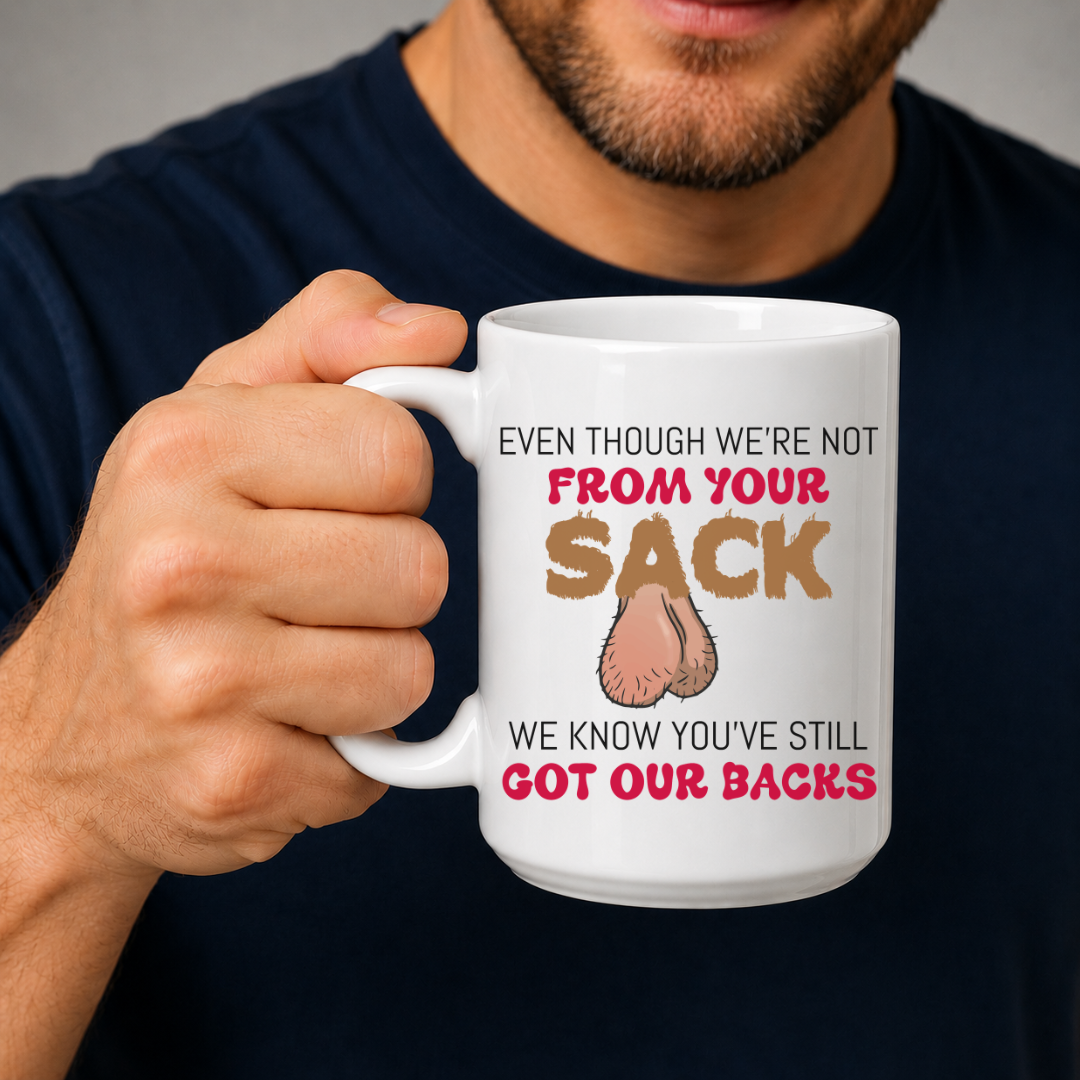 A man in a navy blue t-shirt holds a white ceramic mug with a humorous graphic. The mug features the text 'EVEN THOUGH WE'RE NOT FROM YOUR SACK WE KNOW YOU'VE STILL GOT OUR BACKS' in brown and red lettering, accompanied by a cartoon drawing of a hairy balls. Grab your mug from Coffee and Moo