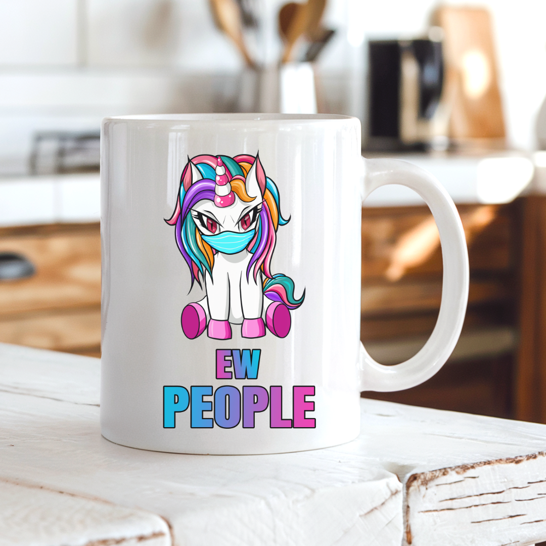 A white ceramic coffee mug with a cartoon unicorn graphic and the text 'EW PEOPLE' printed on it, sitting on a white wooden table in a blurred kitchen setting. from Coffee and Moo.