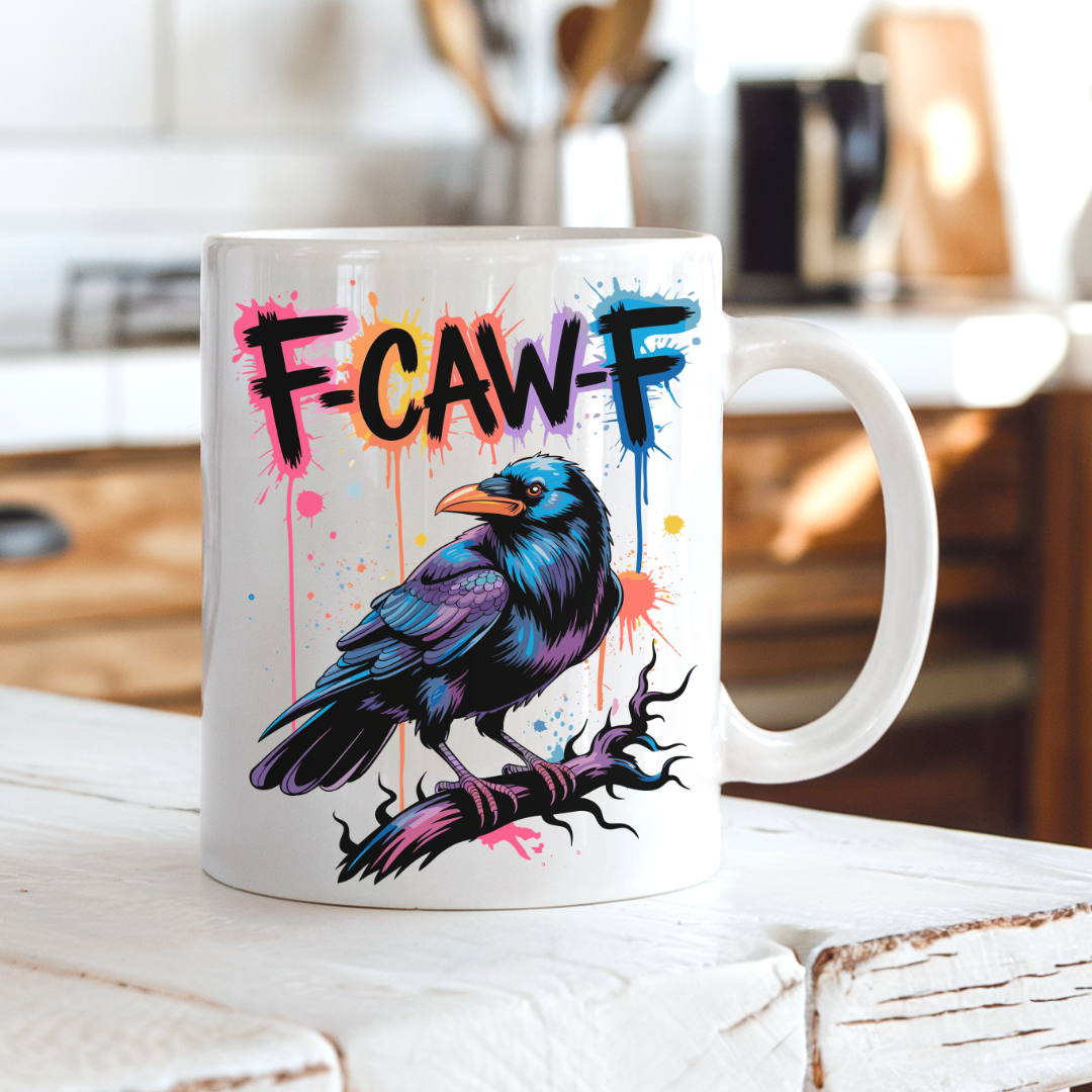 A white ceramic mug with a colorful graphic of a raven perched on a branch under the text 'F-CAWF' in black and blue lettering with paint splatters, set on a rustic white wooden table with a blurred kitchen background.