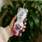 A hand holding a 20oz tumbler decorated with a realistic black and white cow face and pink floral accents, set against a blurred background of green houseplants. Grab yours from Coffee & Moo NZ