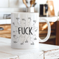A white ceramic coffee mug with a black hand-drawn style graphic spelling out 'FUCK' with other words like 'THEM', 'HER', 'YOU', 'FACE', 'IT', 'HIM', 'THIS', 'OFF', 'WAD', 'THAT', and 'HEAD' arranged around it. The mug is sitting on a white wooden table with a blurred kitchen background. From Coffee and Moo NZ.
