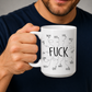 A white ceramic mug with black hand-drawn text and doodles, held by a man wearing a navy blue t-shirt against a neutral gray background. From Coffee and Moo NZ.
