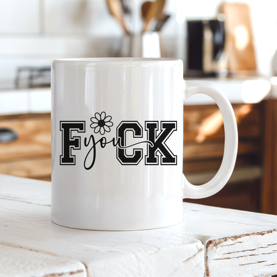A white ceramic coffee mug with the text 'FUCK YOU' printed in black, sitting on a rustic white wooden table with a blurred kitchen background. From Coffee and Moo NZ