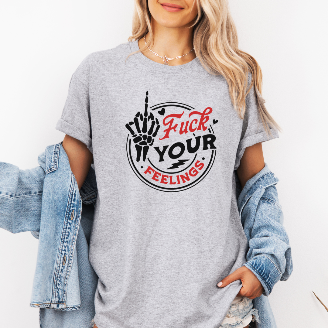 A woman wearing a heather gray graphic t-shirt with a black and red 'Fuck Your Feelings' design, styled with an open light blue denim jacket against a plain white background. Purchase from Coffee and Moo NZ.