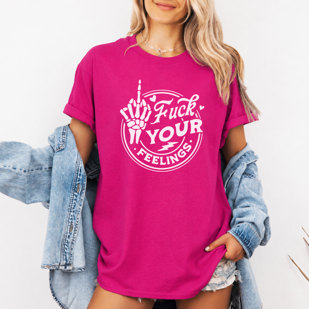 A woman wearing a bright pink graphic t-shirt with a white middle finger hand design and the text 'Fuck YOUR Feelings'. She has a light-wash denim jacket draped off her shoulders and is wearing denim shorts against a plain white background. Purchase from Coffee and Moo NZ.