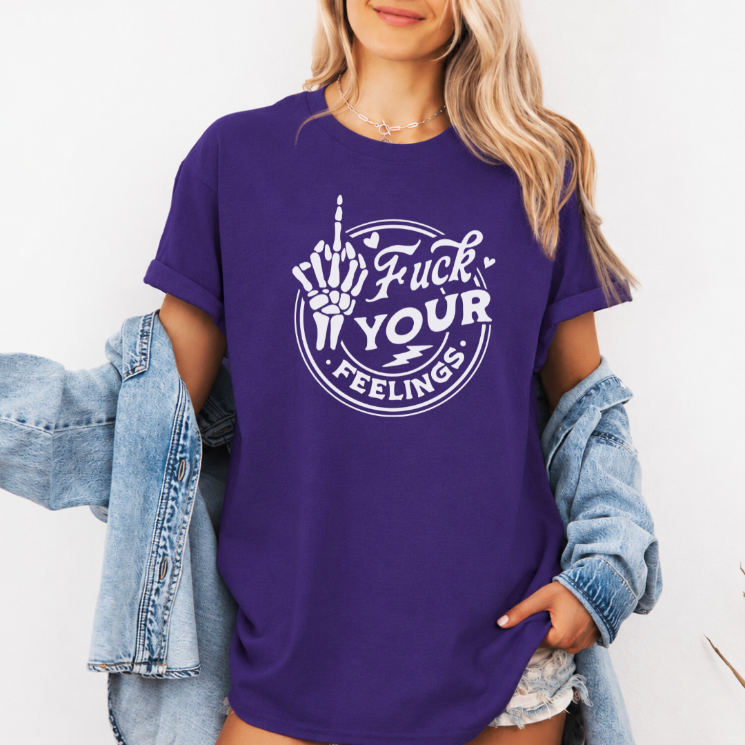 A woman wearing a purple short-sleeve graphic t-shirt with a white circular design that reads 'Fuck YOUR FEELINGS' and shows a middle finger skeleton hand. She has a light blue denim jacket draped off her shoulders and is posing against a plain white wall. Purchase from Coffee and Moo NZ.