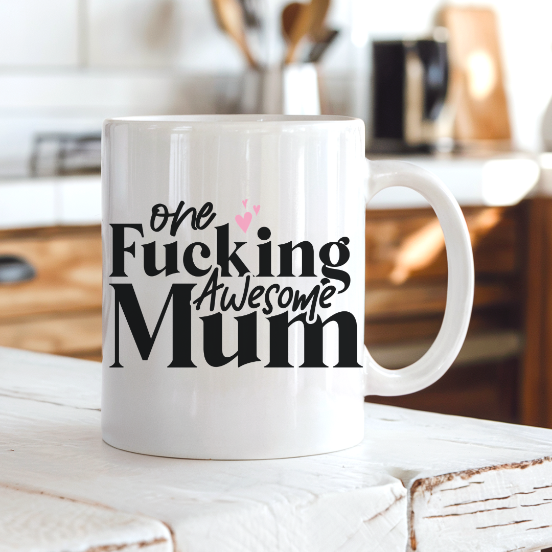 A white ceramic coffee mug with the text 'one Fucking Awesome Mum' in black and pink lettering, sitting on a rustic white wooden table in a blurred kitchen setting. From Coffee and Moo nz