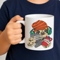 A child holding a white ceramic mug with a cartoon graphic of a sloth wearing an orange beanie, green headphones, and a red shirt while holding a game controller. Grab your gaming sloth mug from Coffee and Moo NZ.