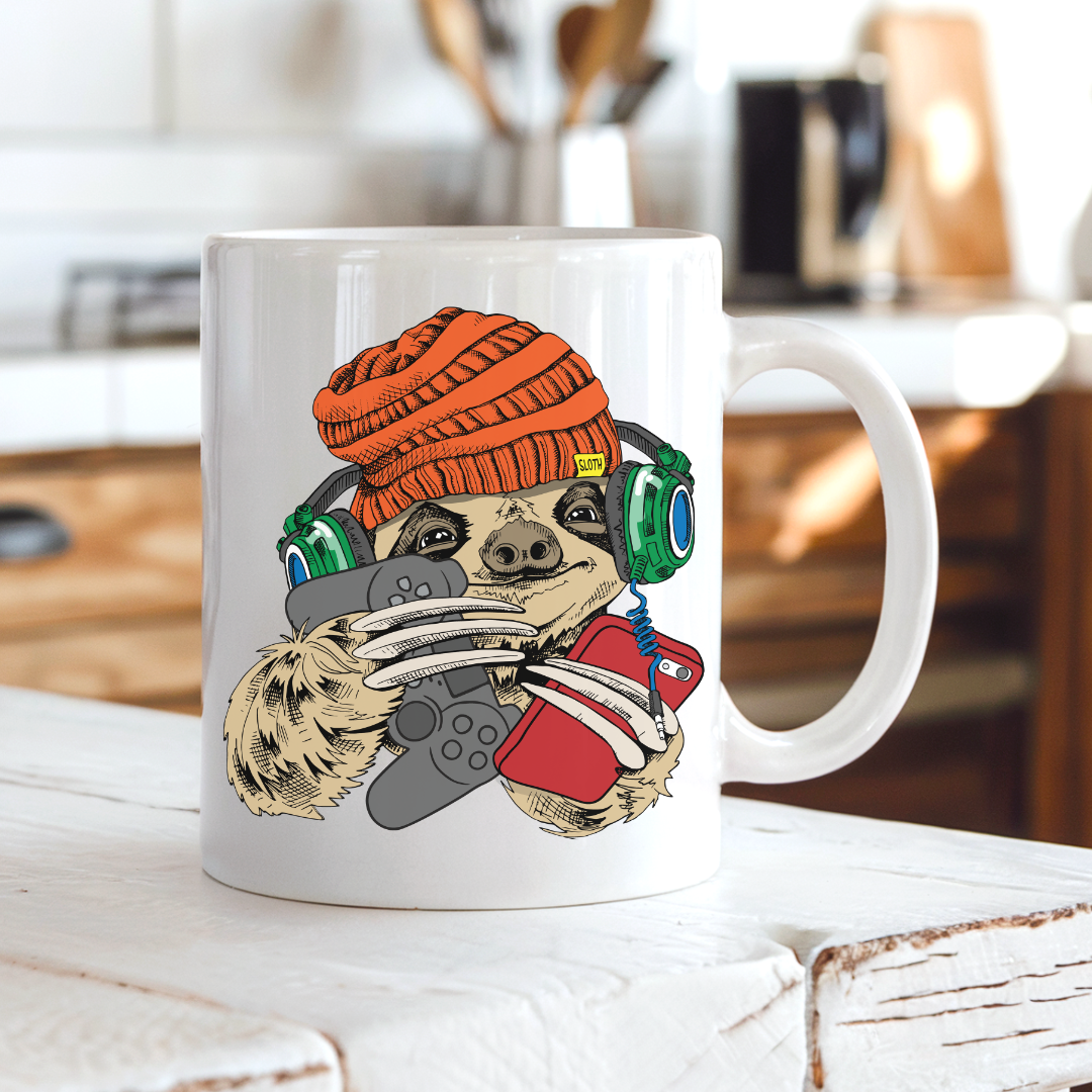 A white ceramic mug with a colorful cartoon sloth graphic wearing an orange beanie, green headphones, and holding a game controller, set on a rustic white wooden table with a blurred kitchen background. Grab your gaming sloth mug from Coffee and Moo NZ.