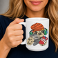 A person holding a white ceramic mug with a colorful cartoon graphic of a sloth wearing an orange knit hat and holding a game controller. The person is wearing a dark navy blue shirt against a neutral grey background. Grab your gaming sloth mug from Coffee and Moo NZ. 
